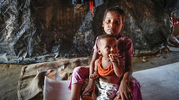 child with younger sibling sitting down inside a makeshift shelter ©Turjoy Chowdhury/Disasters Emergency Committee